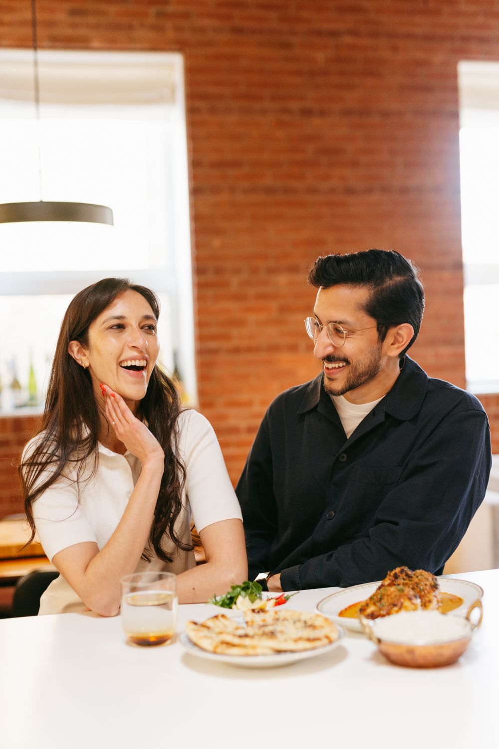 A laughing couple enjoys a meal together at a dining table with Indian food in a modern brick-walled space.
