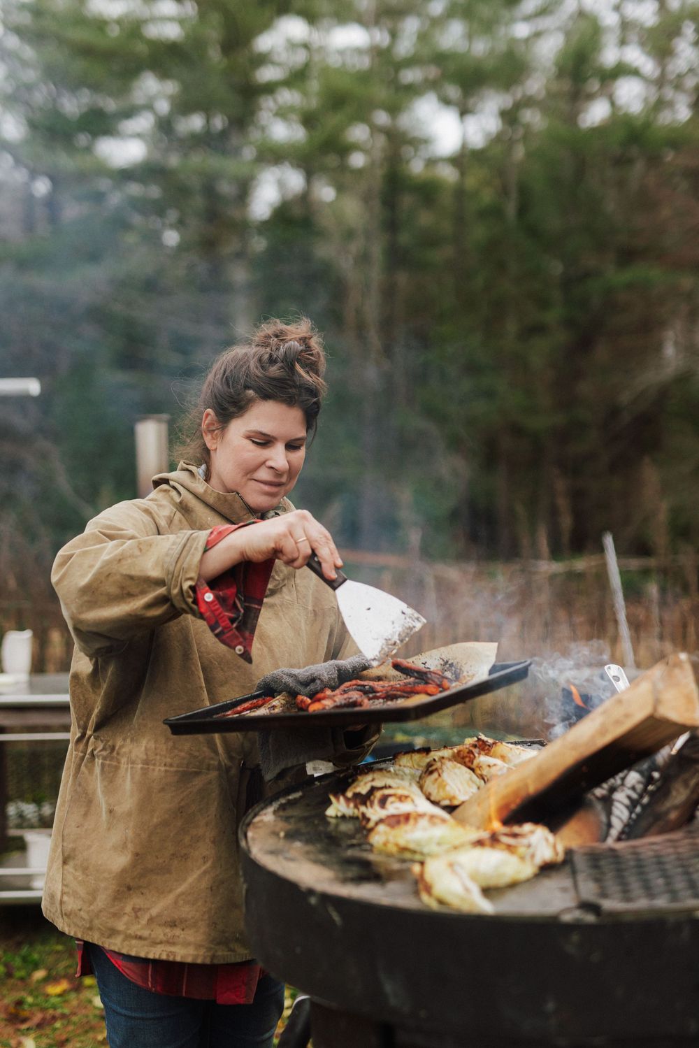 A woman in a tan jacket uses a spatula to tend to vegetables on a large outdoor wood-fire grill.
