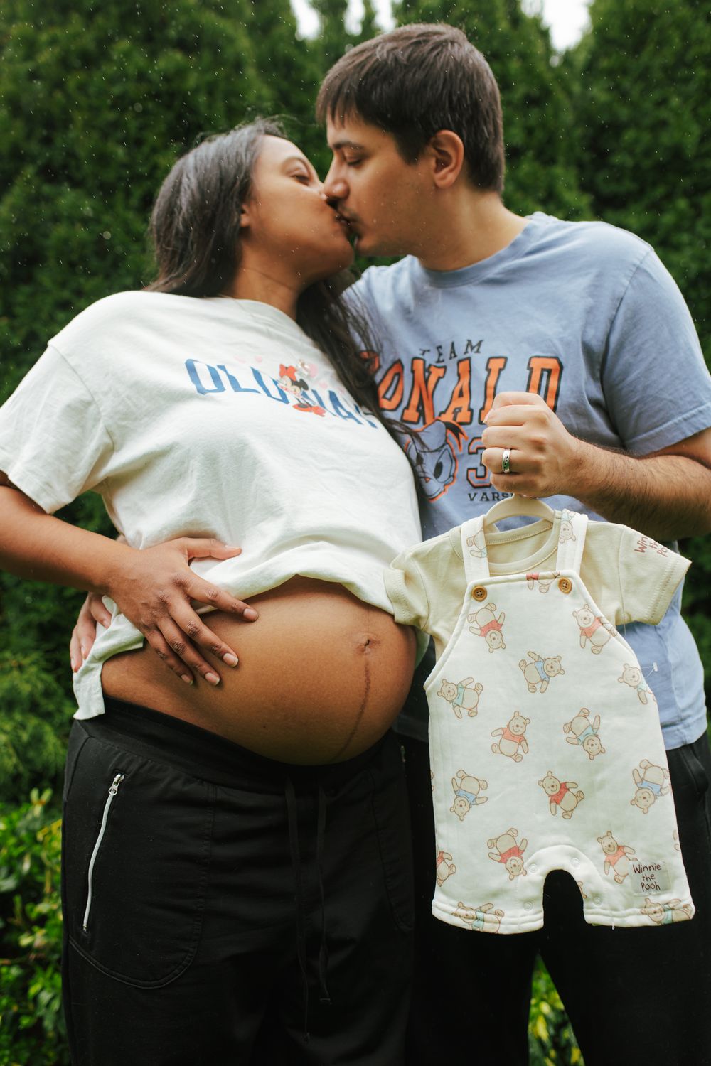 Pregnant couple kissing outdoors while holding a tiny baby romper, celebrating their upcoming arrival together.
