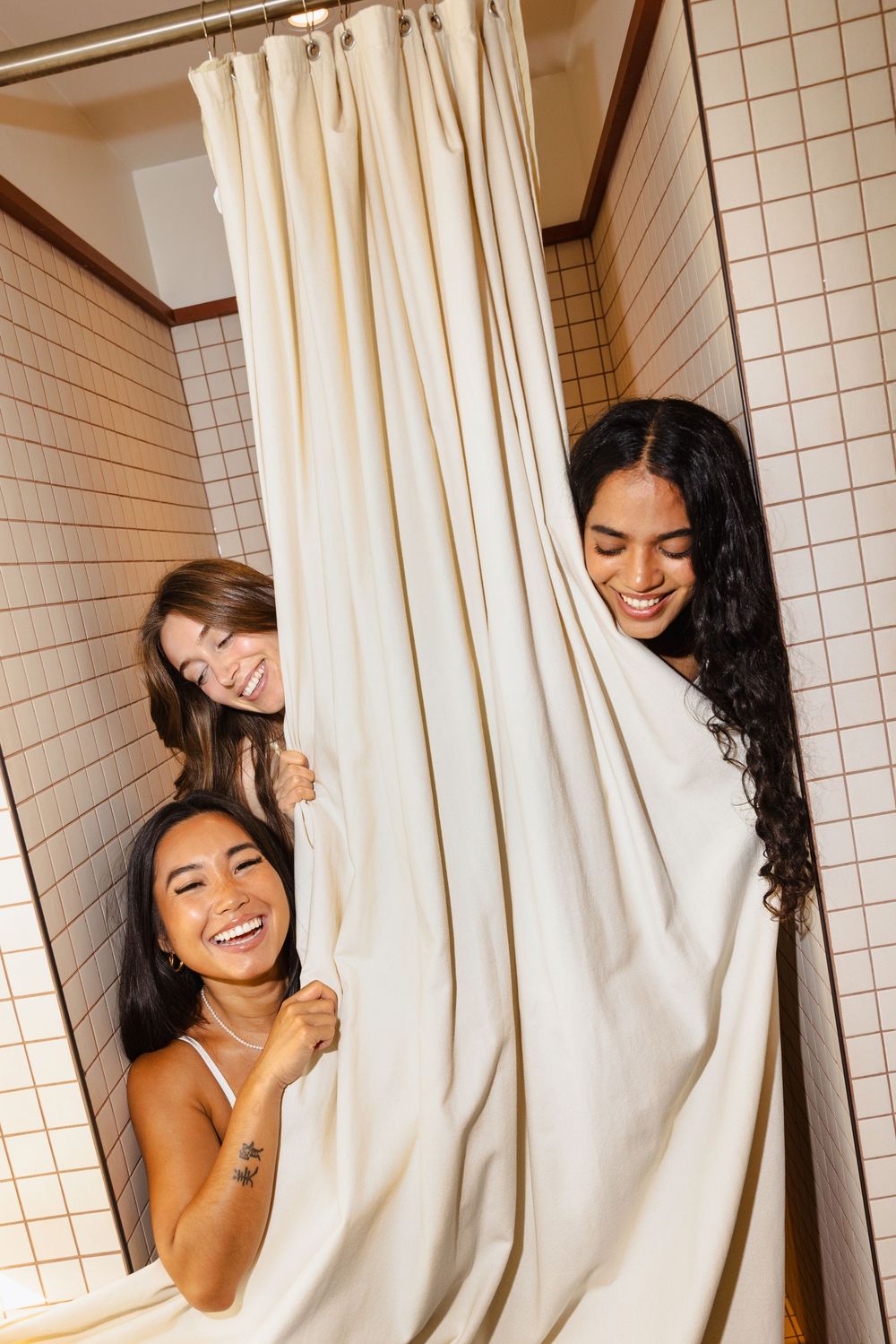 Three smiling women peeking out from behind a white shower curtain in a tiled bathroom.