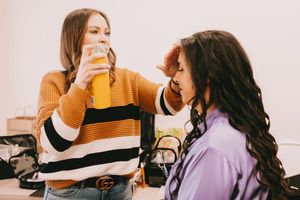 A woman is spraying hair spray on another woman as she covers her eyes.