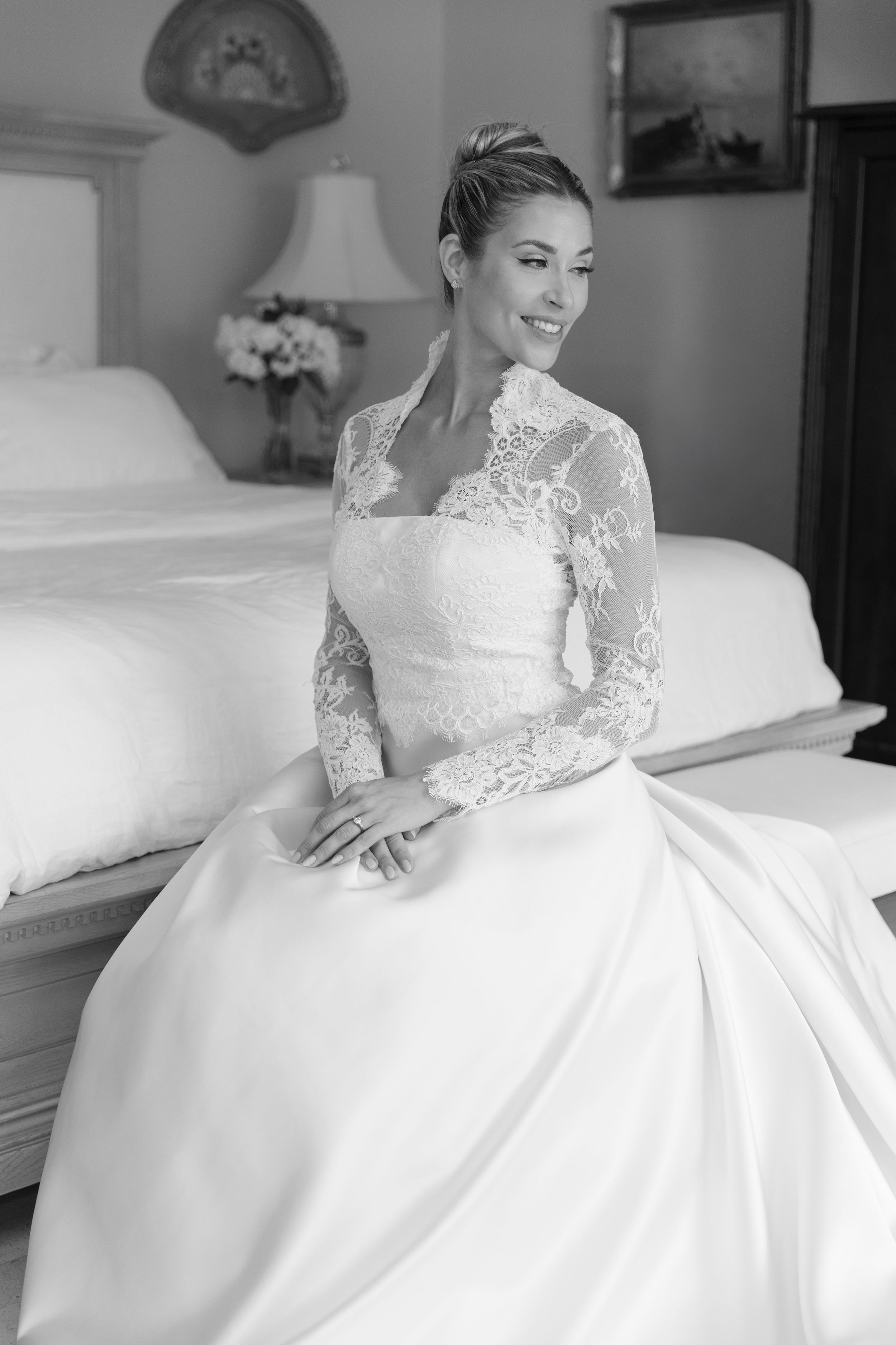 Bride in lace long-sleeve wedding gown sitting on bed, smiling, black and white bridal portrait photo.