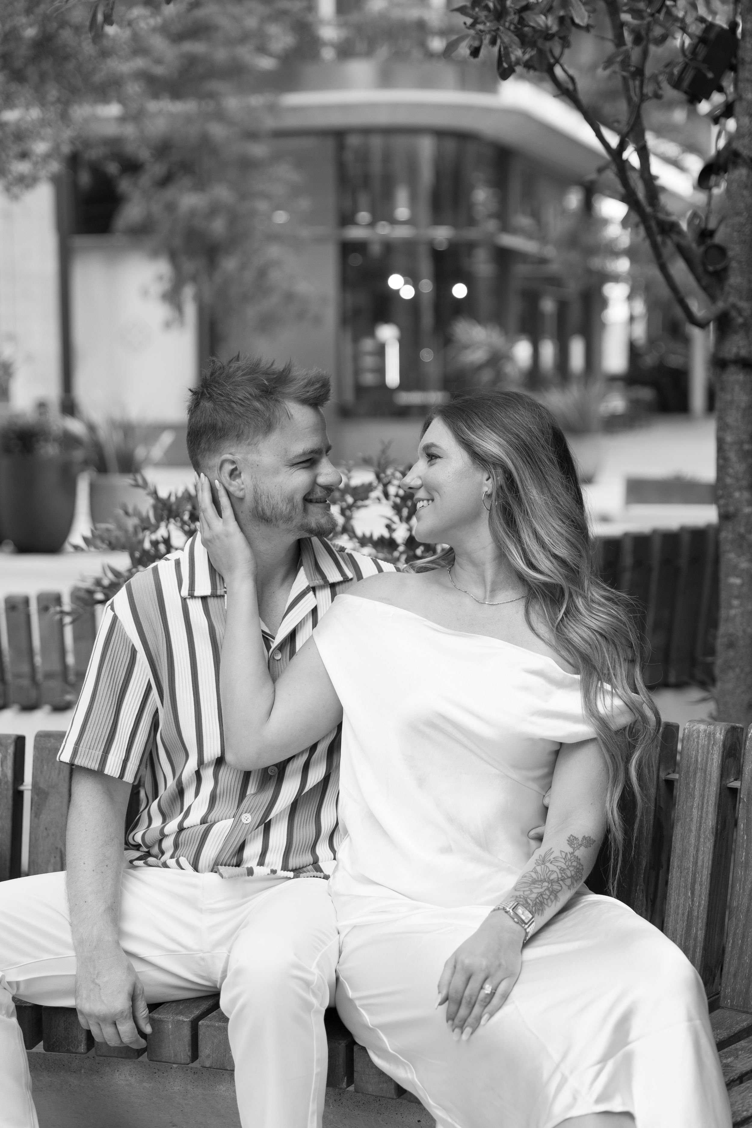 Black and white photo of a smiling couple sitting on a bench outdoors, gazing at each other affectionately.