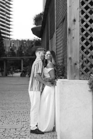 Black and white photo of a romantic couple embracing near a wooden lattice wall on a cobblestone street.