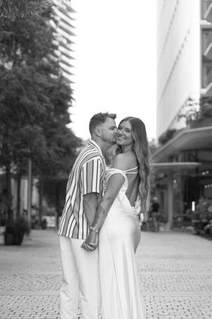 Black and white photo of a couple embracing and kissing on a city street, woman in white dress smiling.