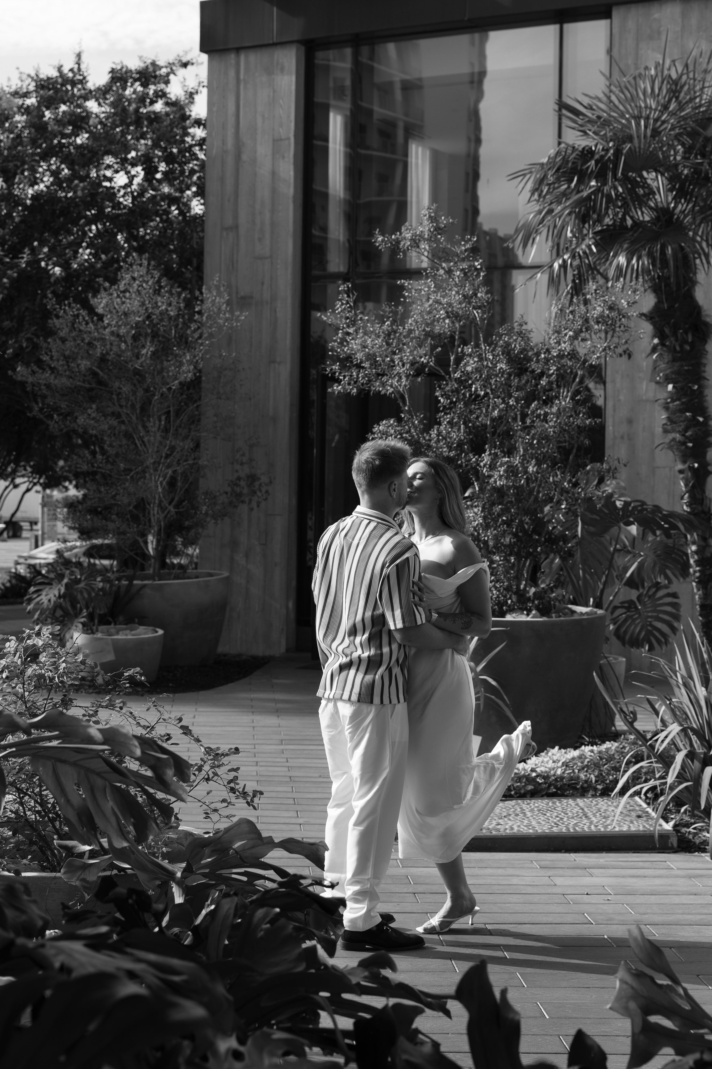 Black and white photo of a romantic couple embracing on a wooden deck surrounded by lush tropical garden plants.