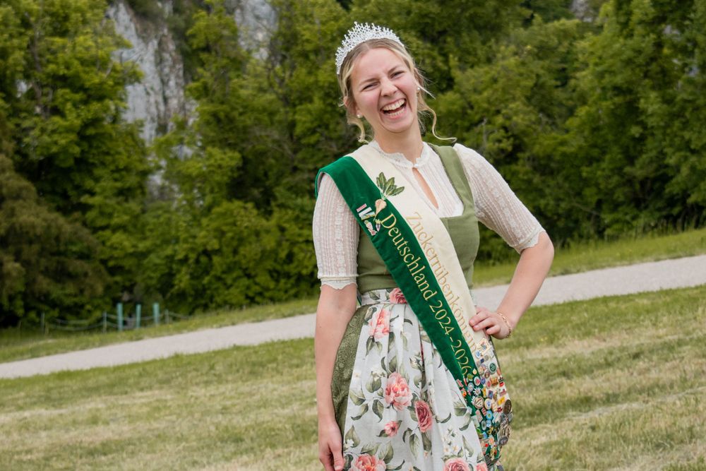 Someone wearing a floral dirndl dress and green sash stands outdoors with a tiara in a scenic natural setting.