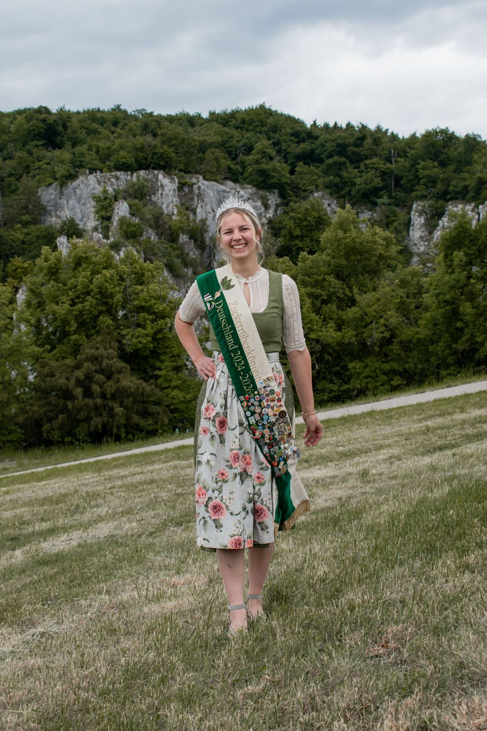 Person wearing traditional Bavarian or Alpine dress with a sash stands on grassy hillside with limestone cliffs in background.