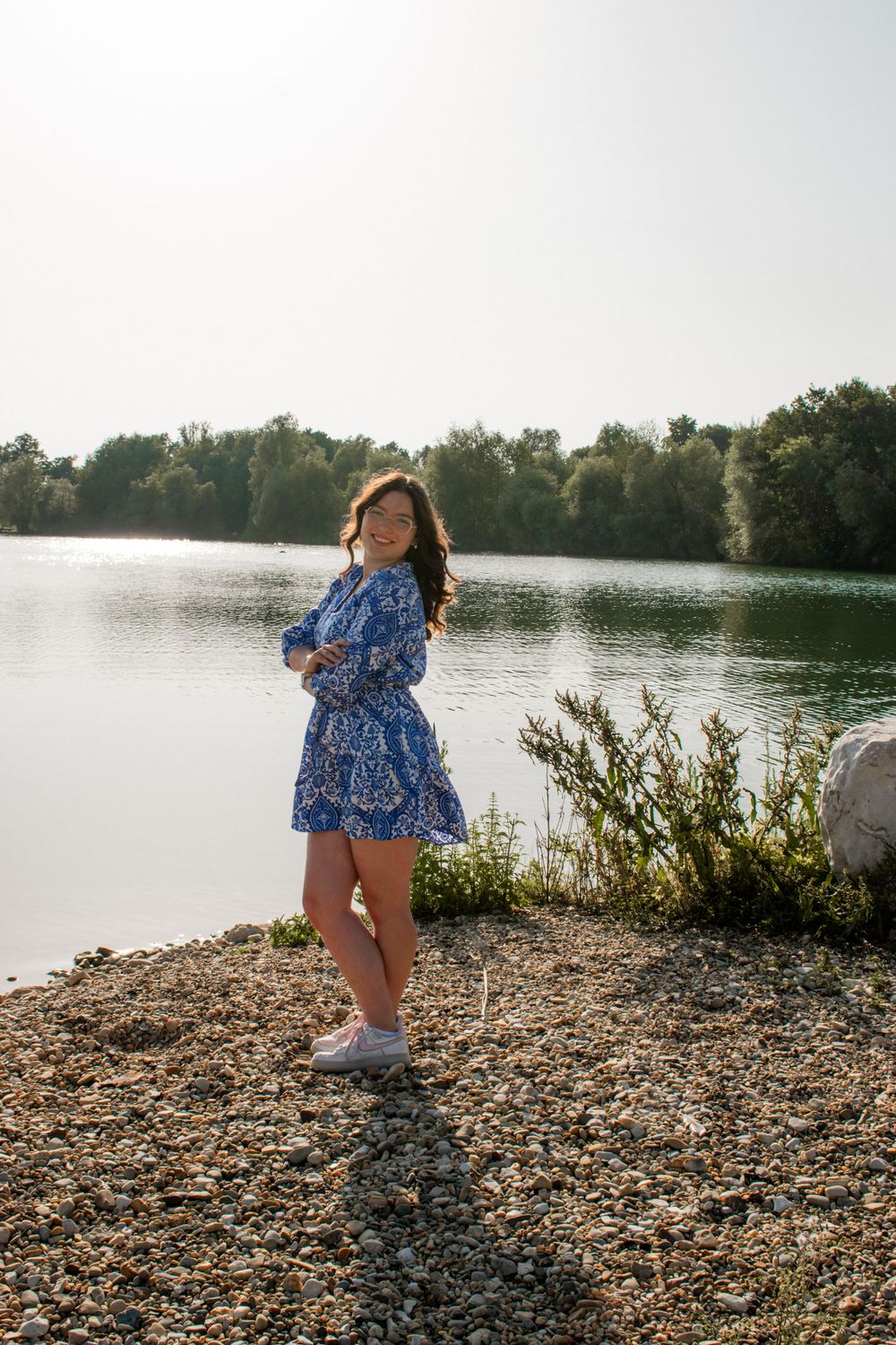 A person in a blue floral dress stands along a rocky lakeshore at sunset with trees in the background.