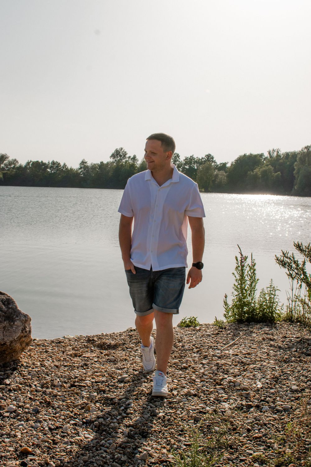 Person in a white shirt and jean shorts walks along a rocky shoreline next to a serene lake during golden hour.