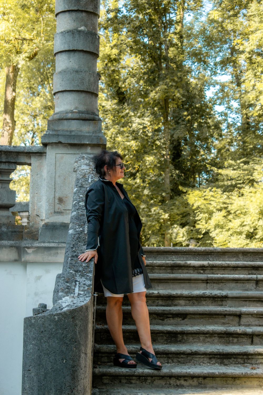 Someone in a dark coat leans against a stone pillar on elegant outdoor stairs surrounded by autumn trees.