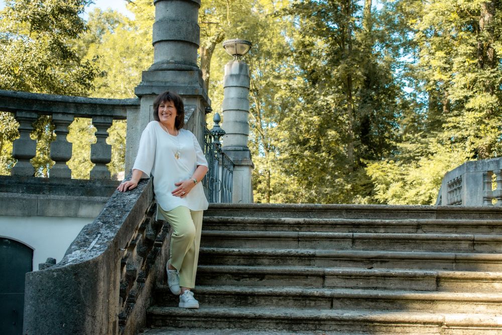 Someone in a casual white outfit stands on ornate stone stairs surrounded by greenery and decorative balusters.