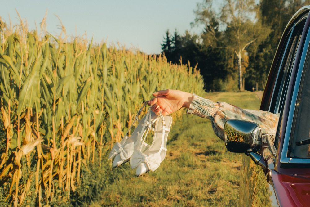 Tattooed hand holding white shoes out of a red car window beside a lush green cornfield on a sunny day.
