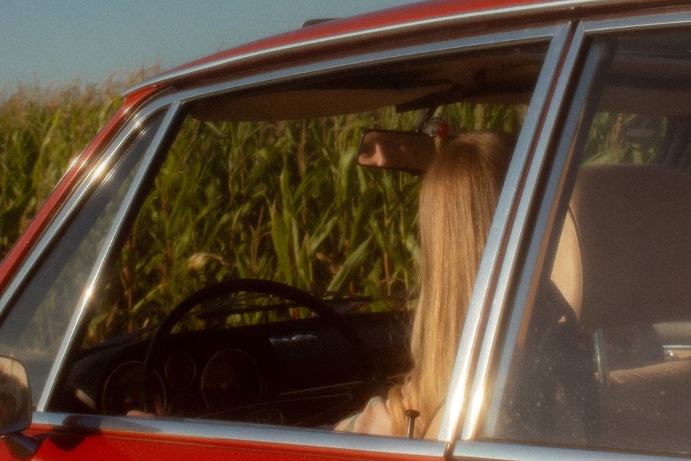 Person with long blonde hair sitting in a vintage red car parked beside a lush green cornfield.