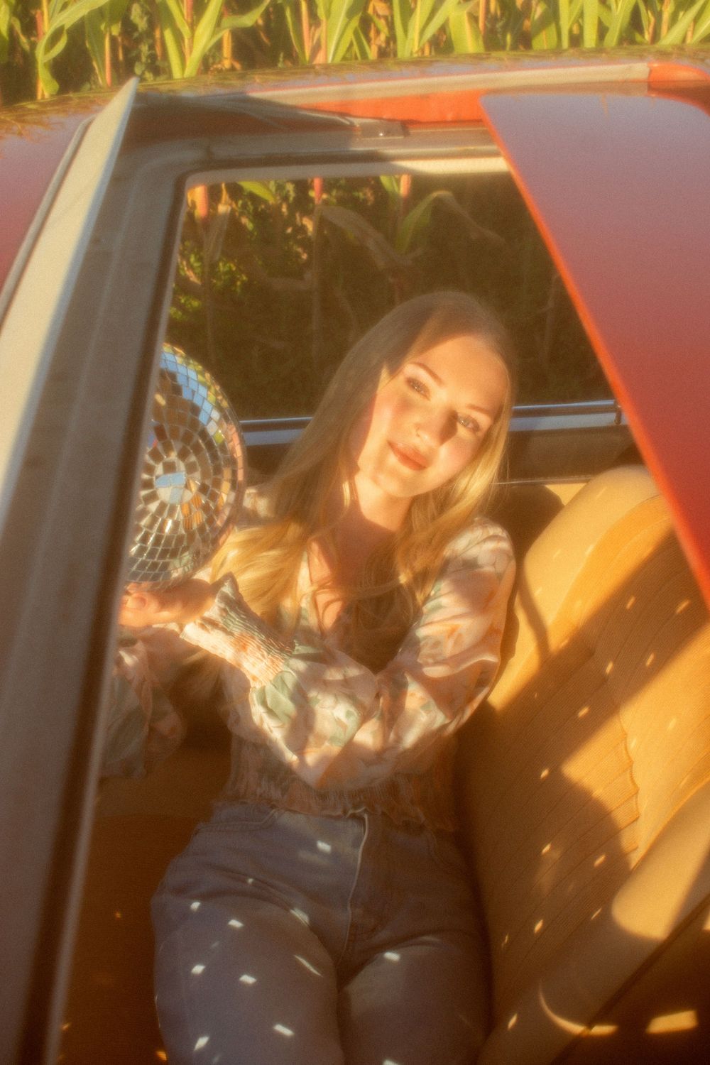 Woman with long blonde hair sitting in a vintage red convertible car near a cornfield in warm golden sunlight.