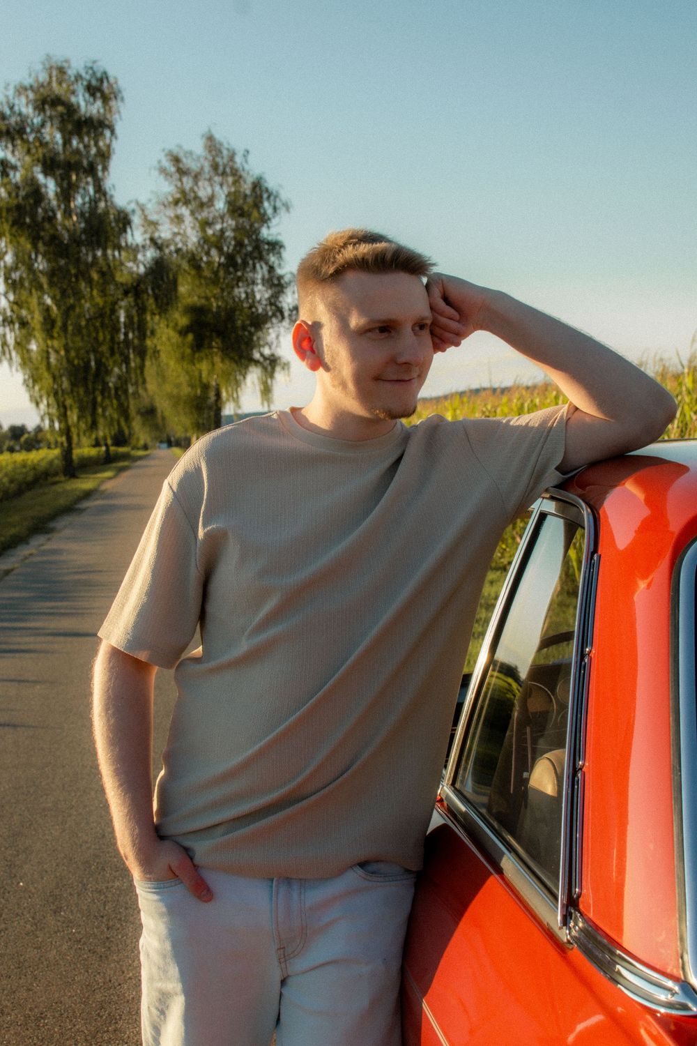 Young man in beige t-shirt leaning on red vintage car on sunny rural road with green trees in background.