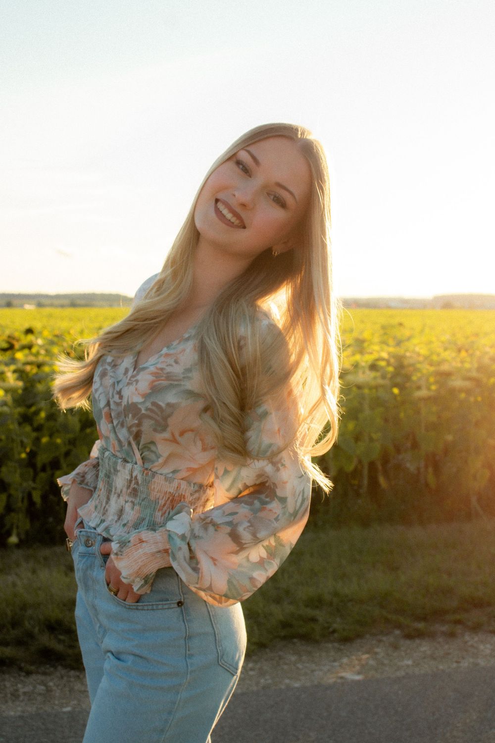 Smiling blonde woman in floral blouse and jeans poses outdoors near sunflower field during golden hour sunset.