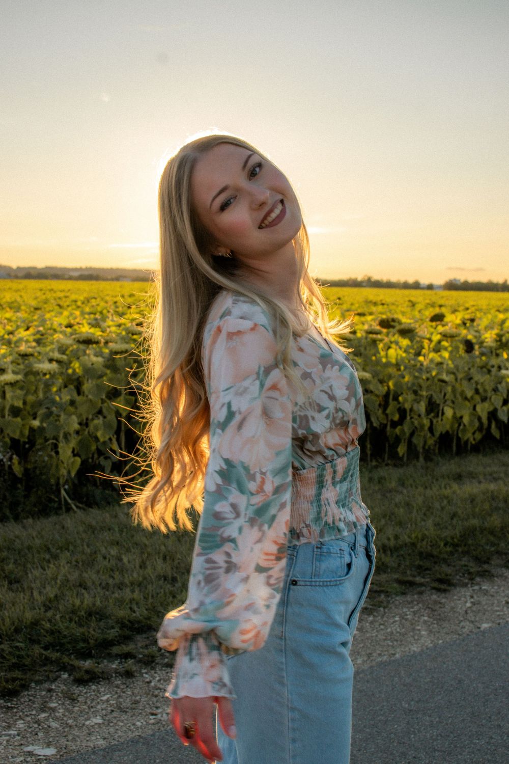 Smiling blonde woman in floral top and jeans poses by sunflower field at golden sunset.