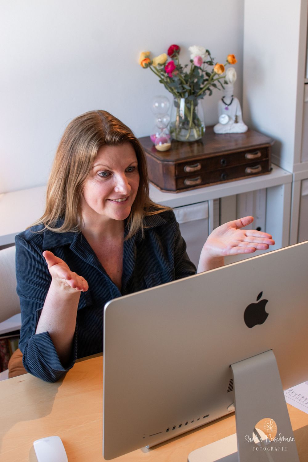 A person in a navy blazer gestures while sitting at a desk with an Apple computer and vase of flowers in the background.