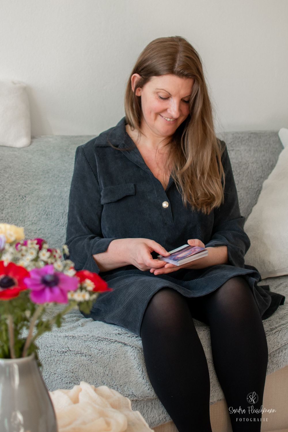 Someone in a black dress sits comfortably on a gray couch while looking at their mobile phone with flowers nearby.