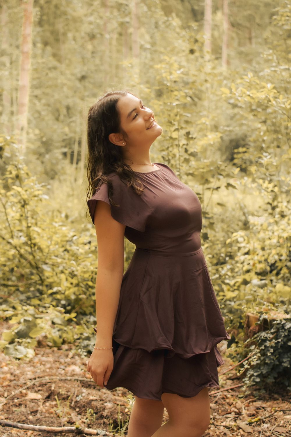 Young woman in brown layered dress poses gracefully in a lush green forest, looking up with a serene smile.