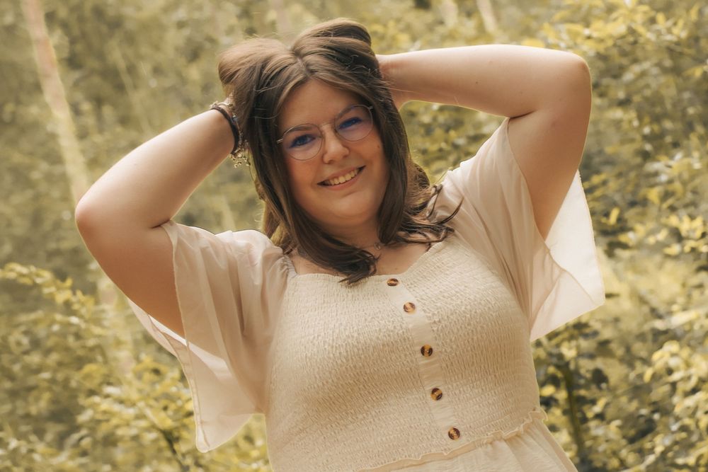 Smiling plus-size woman in cream button-up dress poses outdoors with hands behind head among green trees.