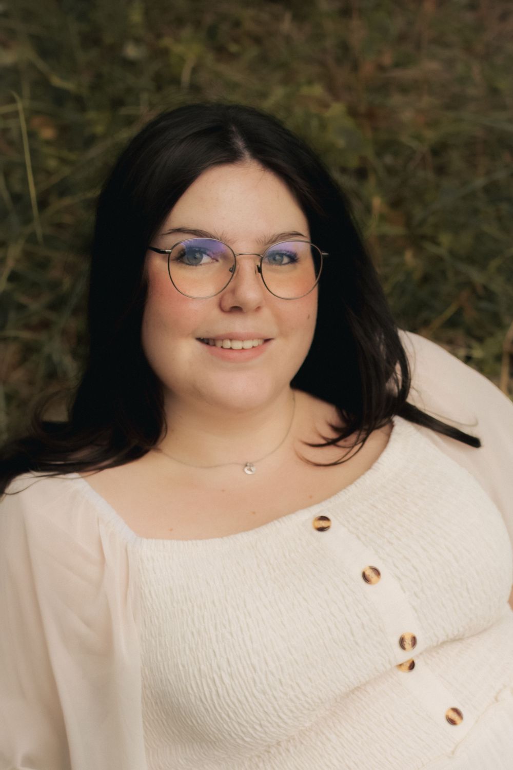 Young woman with dark hair and glasses smiling outdoors, wearing a white top with gold buttons and a delicate necklace.