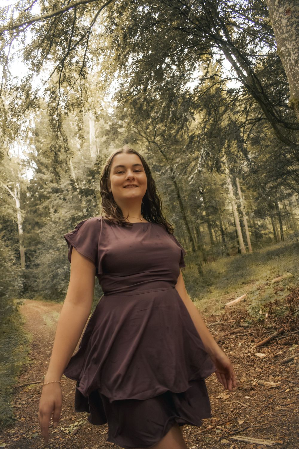 Young woman in purple dress smiling on forest trail surrounded by lush green trees.