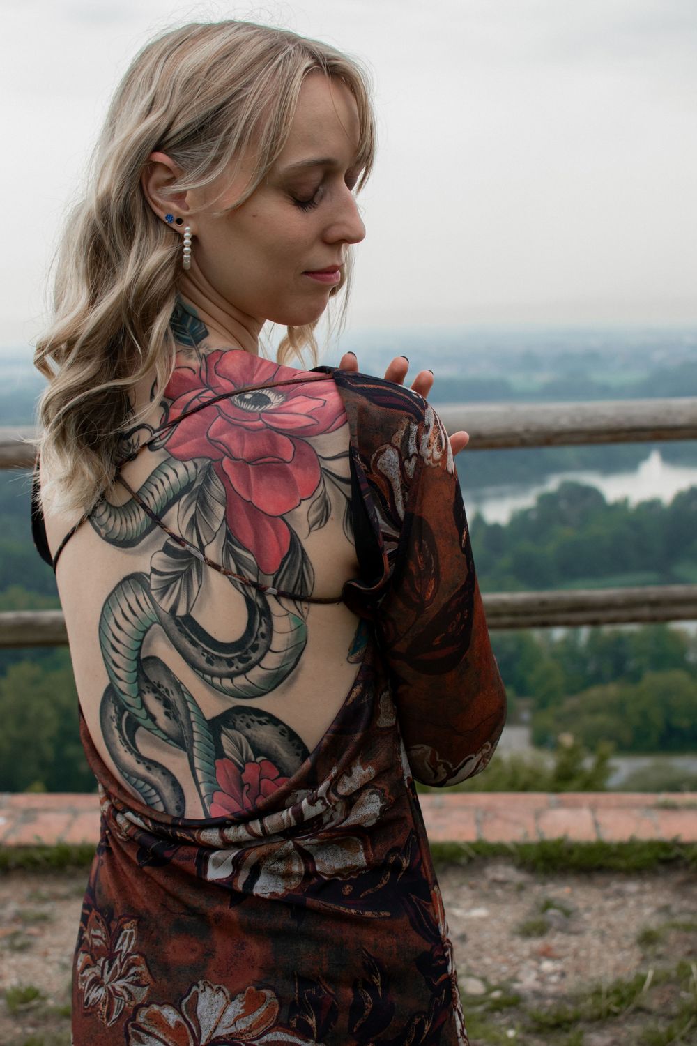 A person with blonde hair in sheer dress shows large floral and snake back tattoo against scenic mountain overlook.