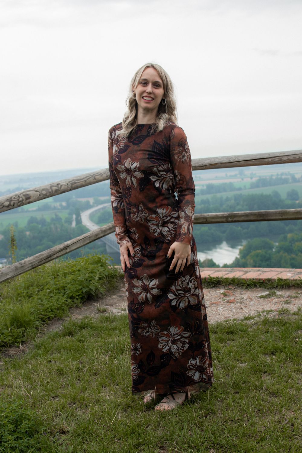 A person in a brown floral dress sits on a grassy overlook with scenic countryside views in the background.