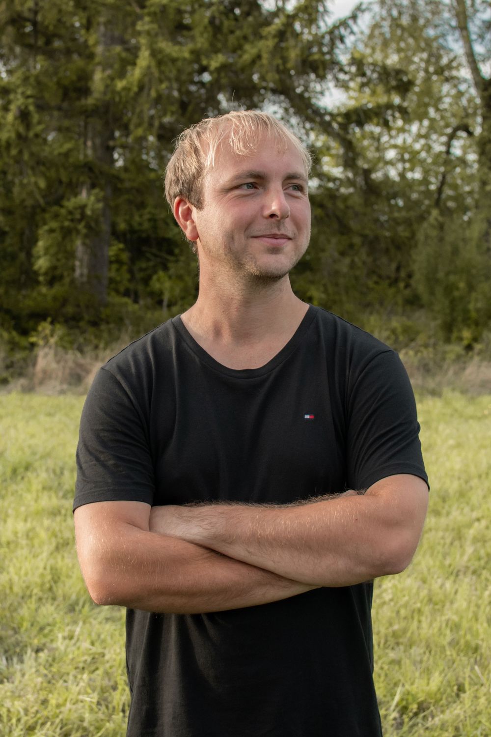 Young man with crossed arms in black t-shirt smiling outdoors with green trees and grass background.