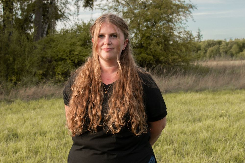 Woman with long curly auburn hair wearing a black shirt standing in a green field with trees behind her.