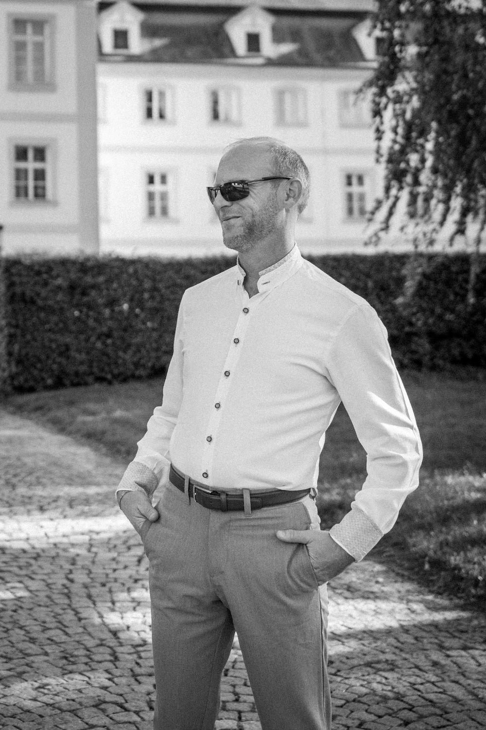 Bald man in white shirt and sunglasses posing confidently outdoors near a historic building in black and white photo.