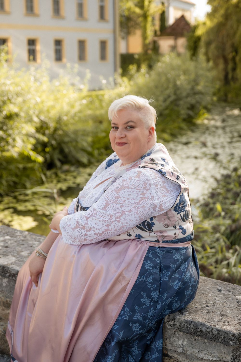 Plus-size woman with short blonde hair in floral dress sits on stone wall in sunny garden.