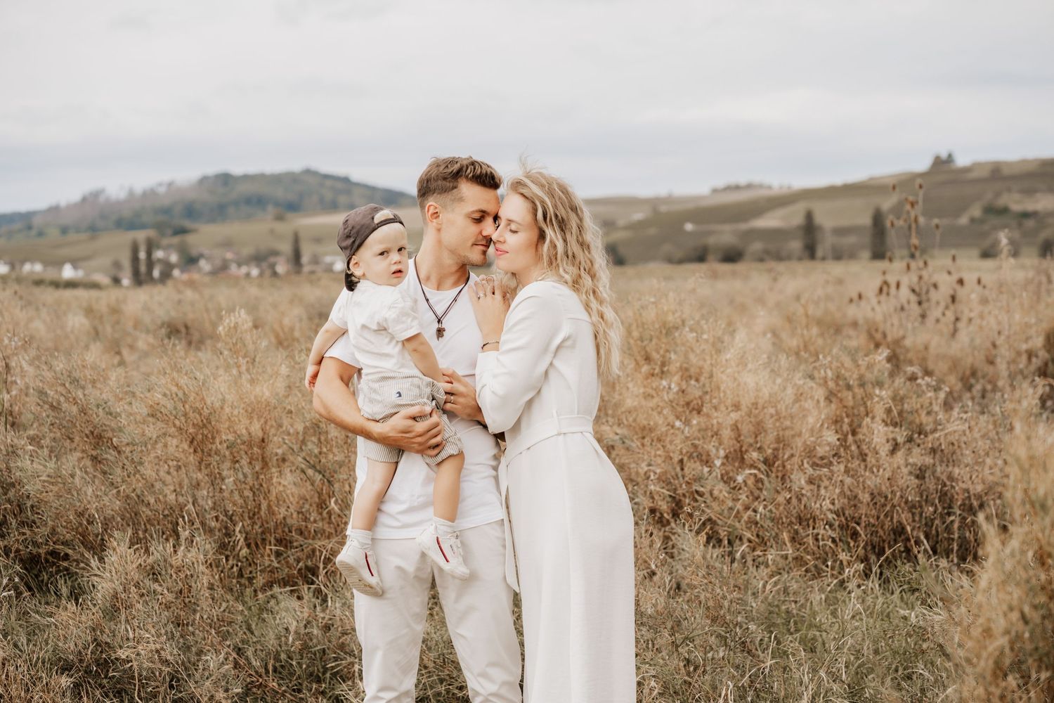 A couple in light colored outfits embraces with their young child in a golden field at sunset.