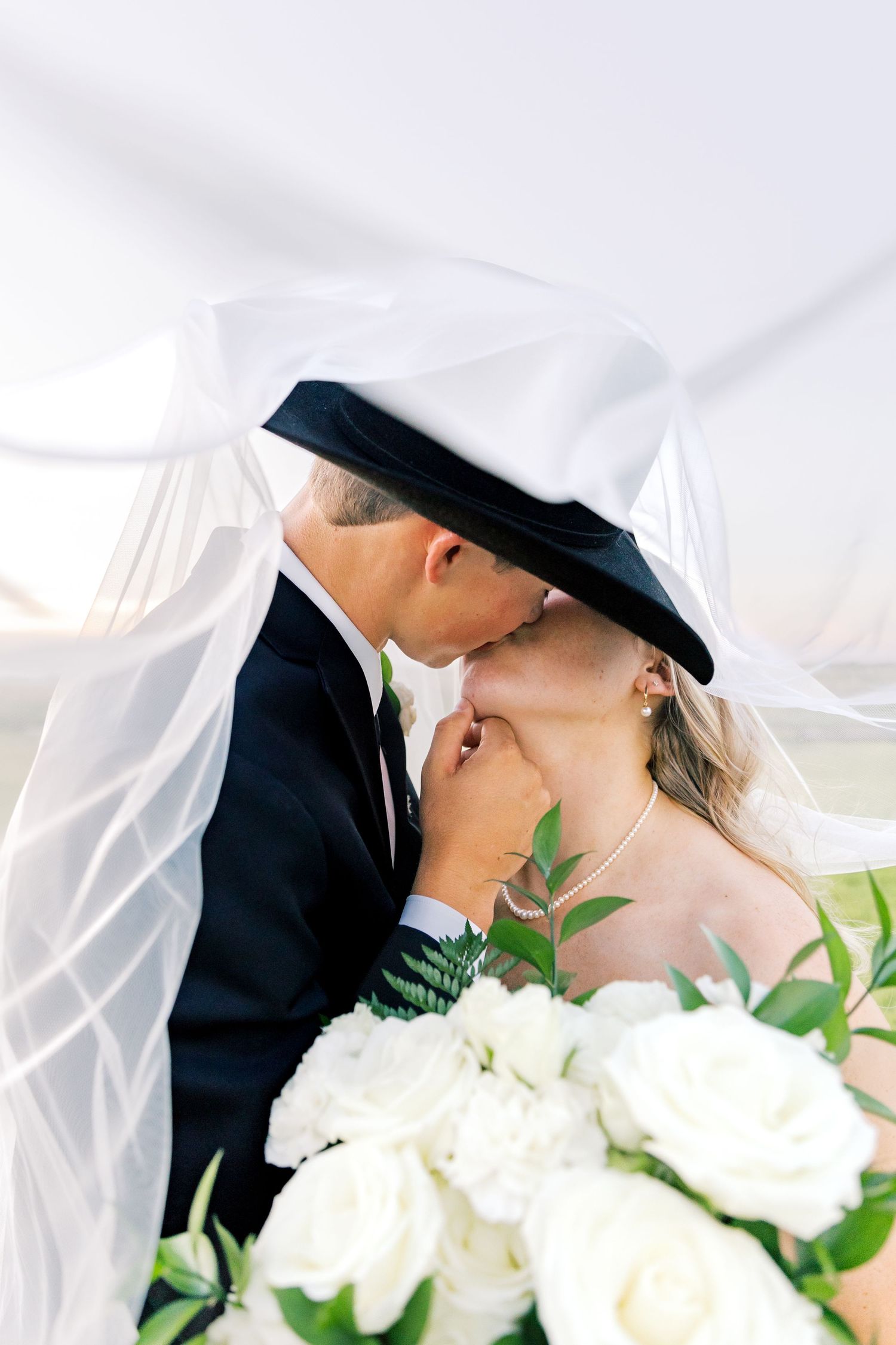 Romantic wedding portrait under flowing white veil with white roses and greenery in foreground.