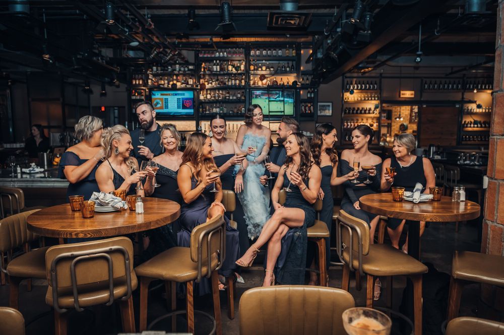 Bride in silver gown celebrated by bridesmaids in black dresses at a lively upscale bar.