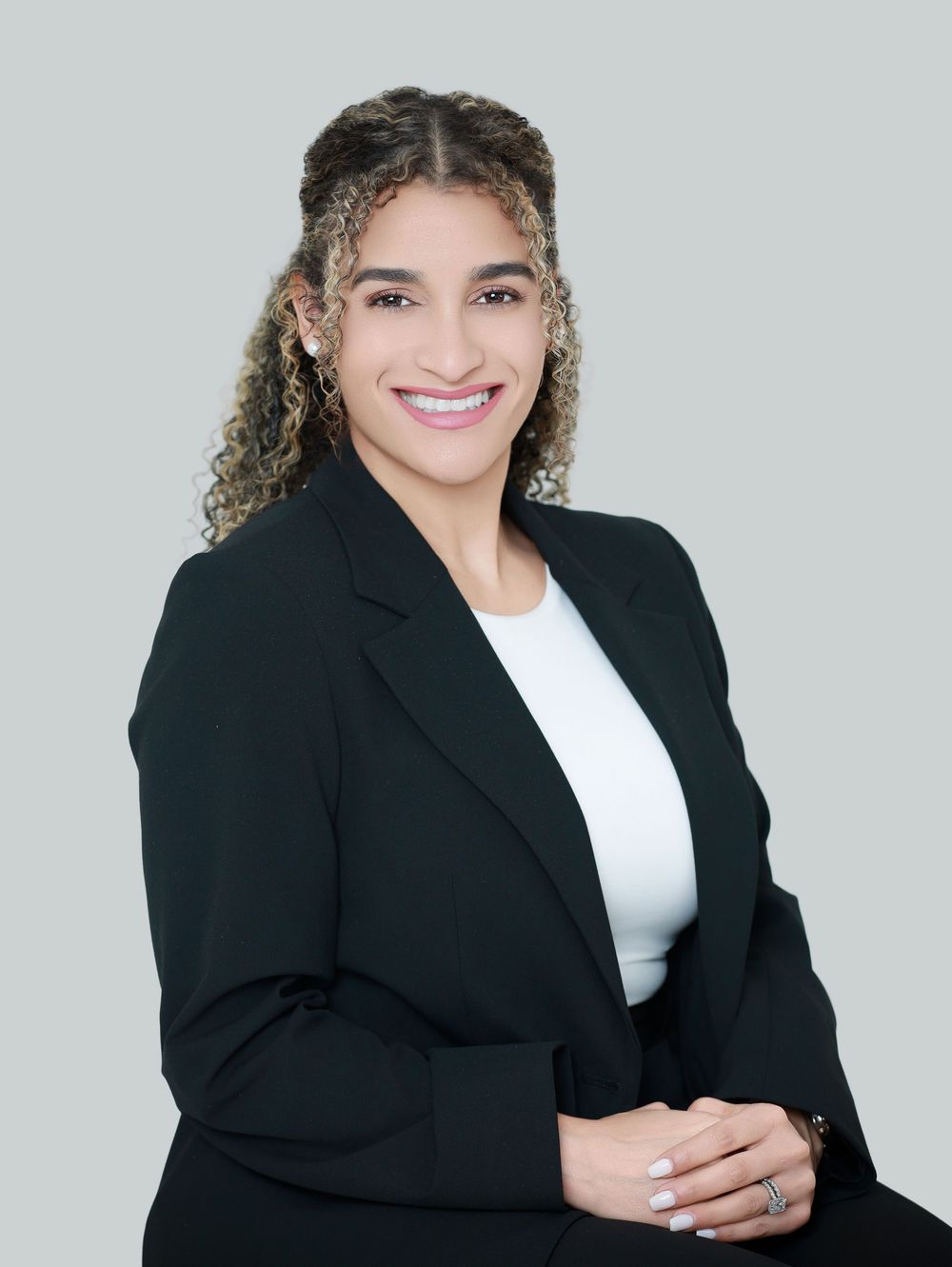 Professional portrait in black blazer and white top against gray background with natural curly hair.