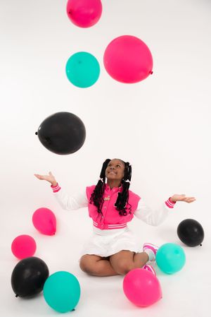 Person lying on floor surrounded by pink, teal and black balloons against white background.