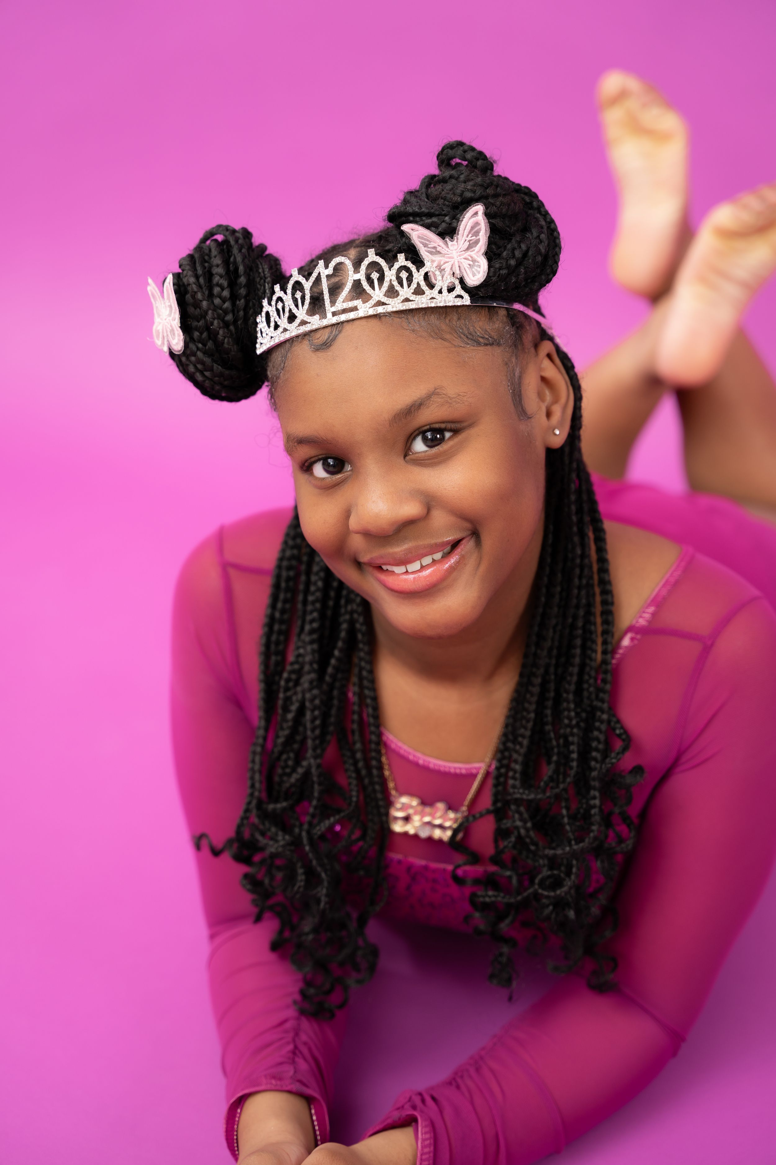 Young dancer in purple leotard with braided hair and tiara poses expressively against pink background.