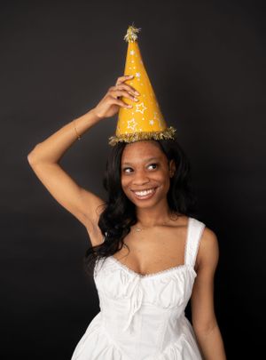 Studio portrait wearing white dress and yellow party hat showing playful expression.