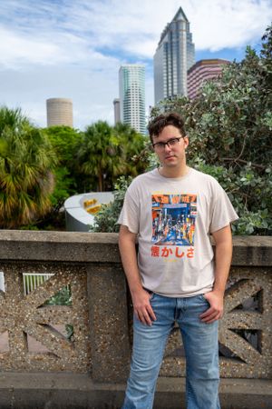 Person wearing graphic t-shirt poses against stone railing with palm trees and city skyline behind.