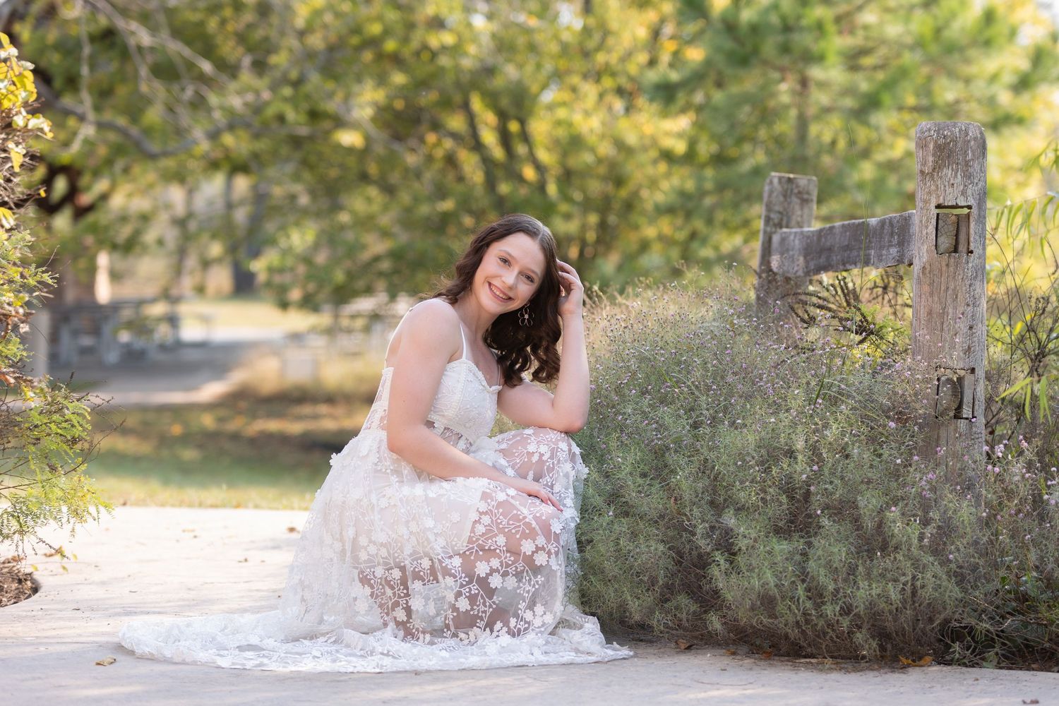 A woman in a white lace dress sits outdoors by a rustic fence surrounded by soft natural lighting and greenery.