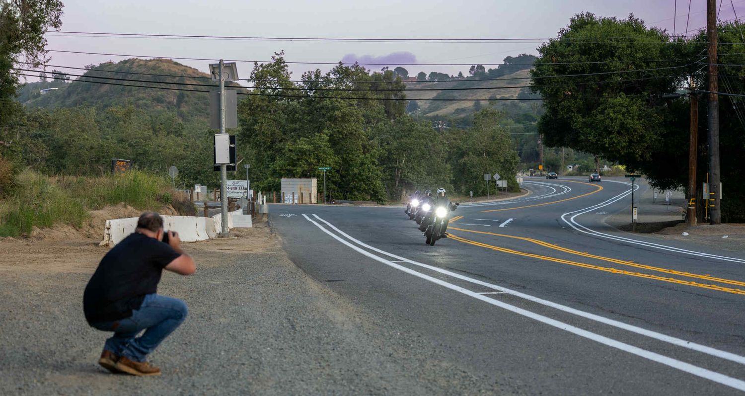 Photographer crouching to take picture of approaching motorcycle on curved road.