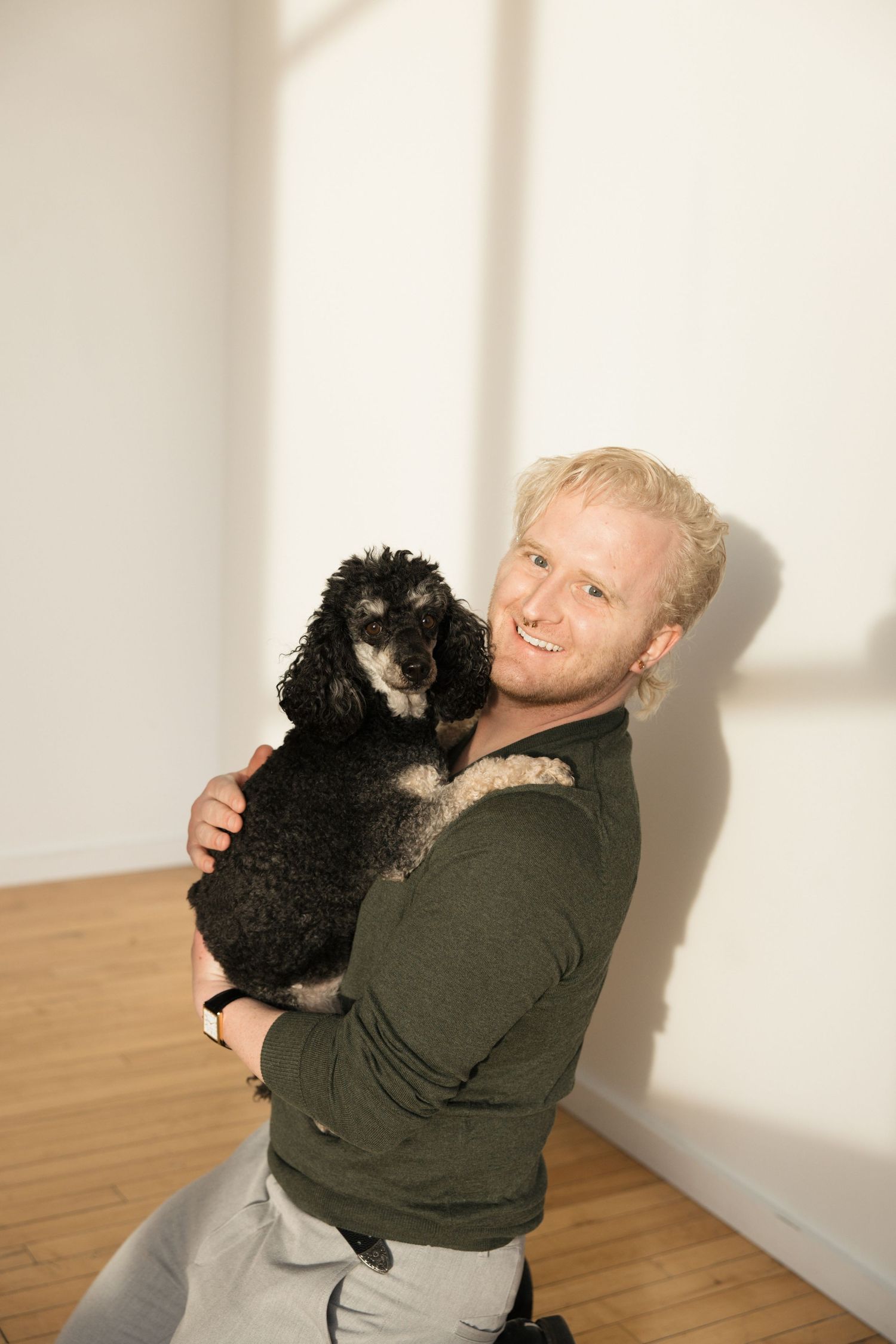 Smiling blond man holding a black and white poodle against a white wall indoors.