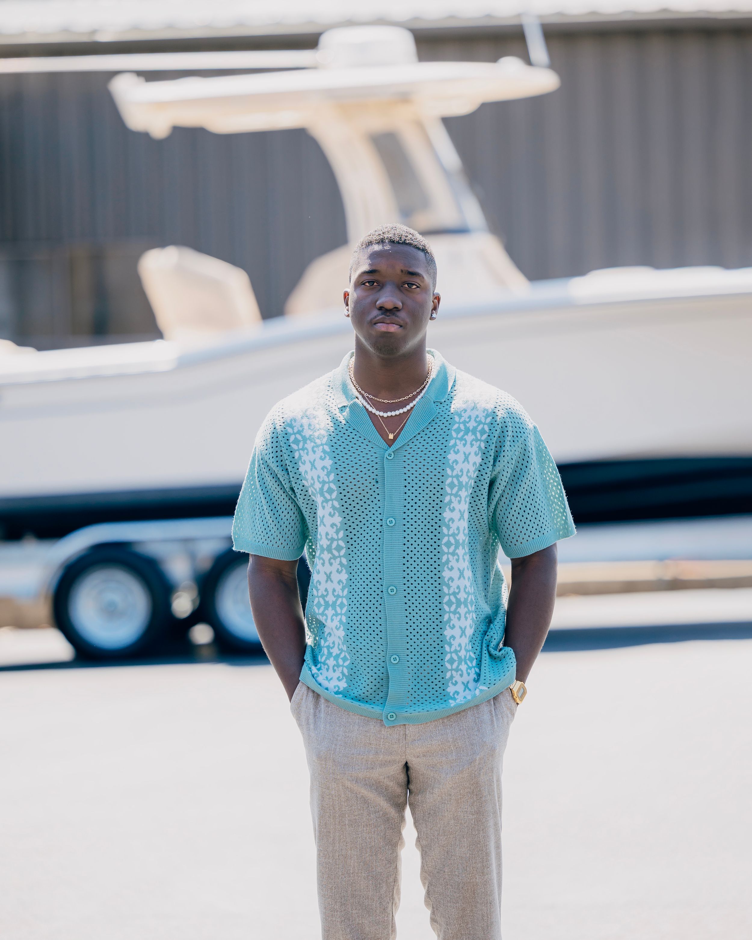 Young man in teal patterned shirt and beige pants stands near boat wearing pearl necklace.