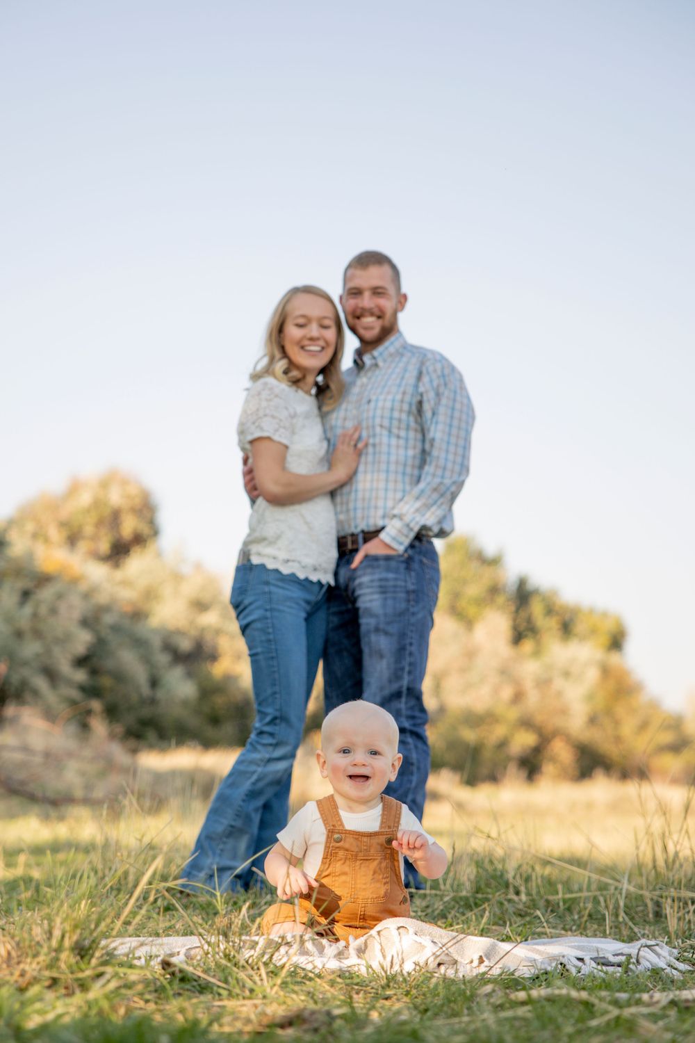 Family portrait in a sunny field with baby sitting in brown overalls.