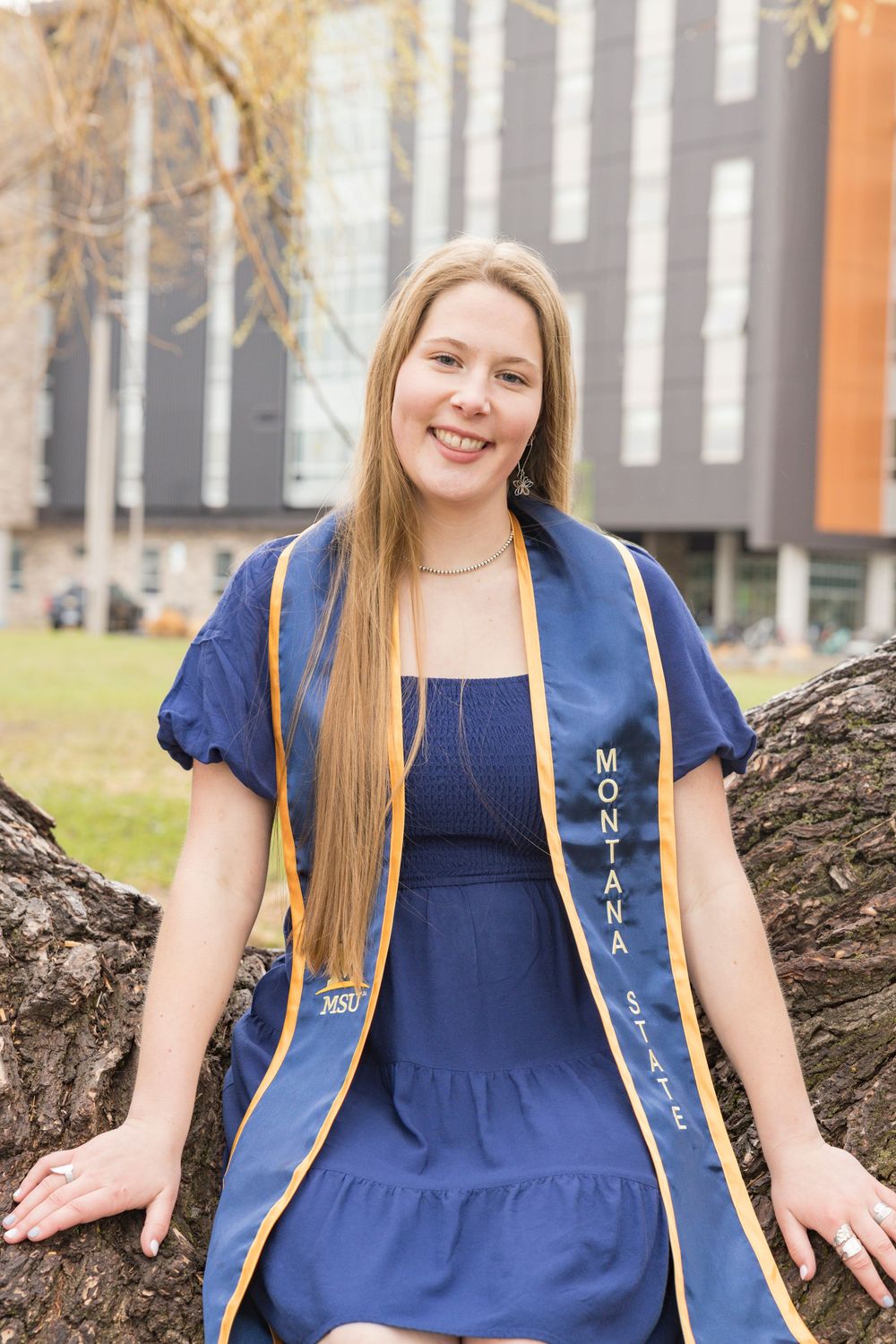 Student wearing blue dress and Montana FFA jacket posing outdoors near tree.