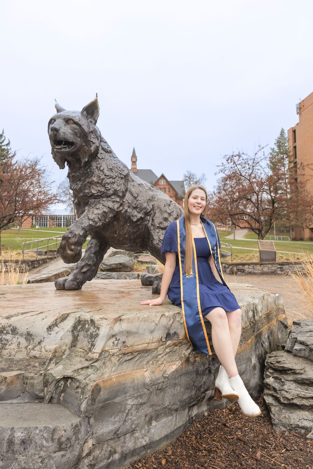 Student poses next to a large metallic husky dog statue on a rocky campus landmark.