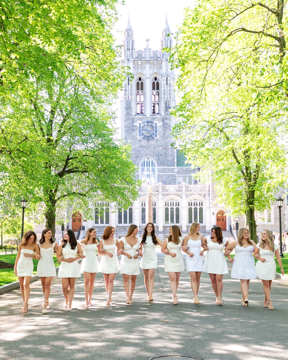 A line of bridesmaids in short white dresses pose together on a pathway in front of a Gothic church with green trees.